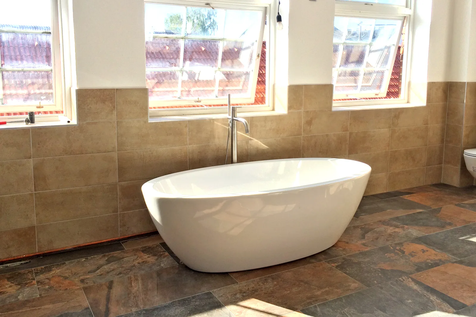 Freistehende weiße Badewanne mit Standarmatur unter drei Dachfenstern, Wandfliesen in warmem Beige und Boden in Schieferoptik