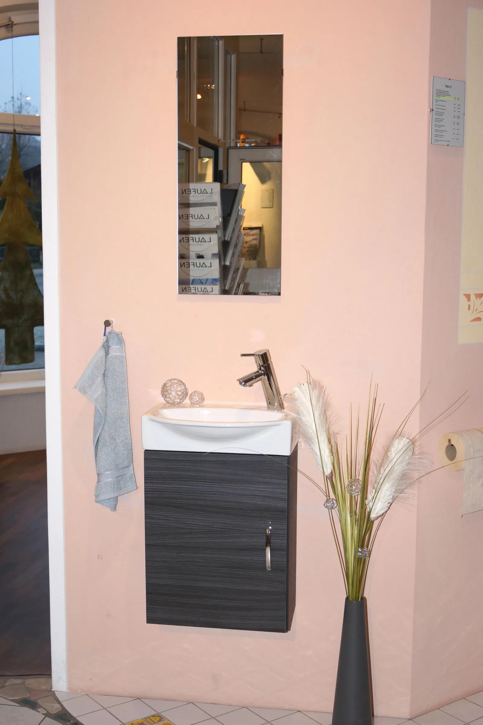 Kleiner Waschplatz im Schauraum mit dunklem Unterschrank, Spiegel, Handtuch und Deko-Gräsern